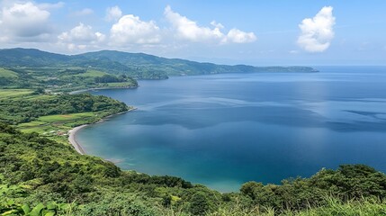 Obraz premium Panoramic coastal view serene blue ocean, lush green hills, and a small secluded bay under a partly cloudy sky