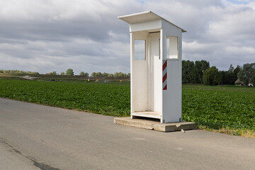 An isolated white booth, a guard shack, stands by a rural road next to a vibrant green field under a cloudy sky.