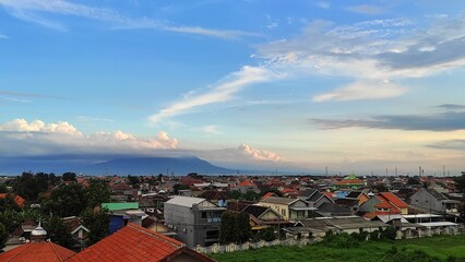 Fototapeta premium Wide-angle cityscape with rows of rooftops in the foreground, a towering volcano partially obscured by clouds in the background, captured during golden hour with soft light and pastel sky tones