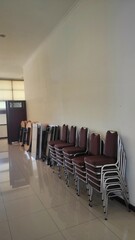 Stacks of brown cushioned chairs and folded tables are neatly arranged against a clean wall in a well-lit, glossy-tiled room, ready for future use.