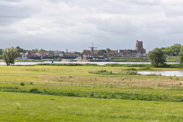 A beautiful panoramic view of the historic town of Woudrichem, Netherlands, featuring its iconic church and windmill, nestled by a wide river and lush green fields. A classic Dutch landscape.