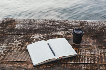 A grid notebook with a pen lies open on a rustic wooden table by the water, next to a black takeaway coffee cup a peaceful setting for writing or reflection outdoors.