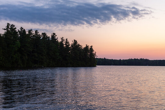 Beautiful sunrise over Lake Lovering, with blue cloud in pink sky and dark trees reflected in choppy water, Magog, Quebec, Canada