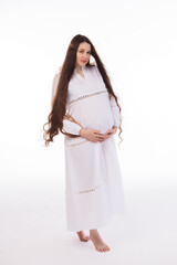 Pregnant woman in white long linen dress with very long hair on white background. Dreamy tender girl expecting a child. Portrait of beautiful pregnant woman in white shirt, smile.