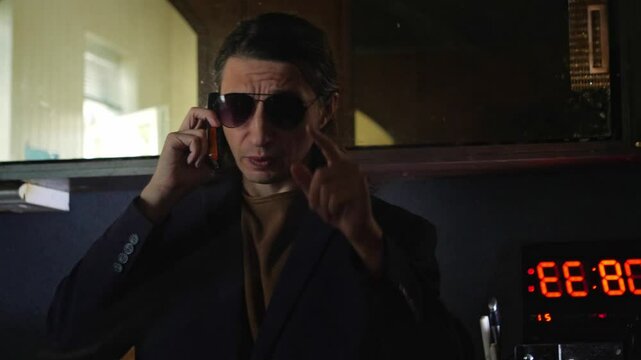 A serious and discreet mood is captured as a man in a suit and sunglasses talks on a mobile phone, with a digital clock in the background. This shot is ideal for depicting clandestine conversations