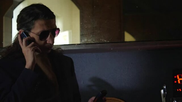 A serious and discreet mood is captured as a man in a suit and sunglasses talks on a mobile phone, with a digital clock in the background. This shot is ideal for depicting clandestine conversations