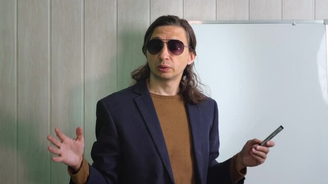 An authoritative and engaging mood is captured as a man in sunglasses and a suit jacket, holding a marker, gestures expressively while standing next to a whiteboard