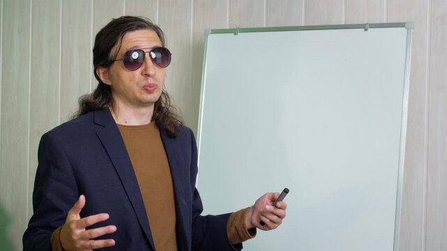 An authoritative and engaging mood is captured as a man in sunglasses and a suit jacket, holding a marker, gestures expressively while standing next to a whiteboard