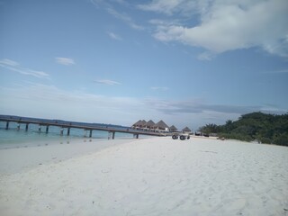 Fototapeta premium Soft white sands stretch beneath a blue sky, with a long pier reaching out to overwater bungalows. The distant tree line and scattered clouds enhance the relaxed island atmosphere