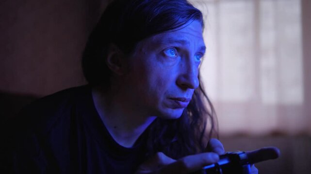 An intensely focused and absorbed mood is captured as a man, illuminated by the blue glow of a screen, plays a video game with a controller in a dimly lit room