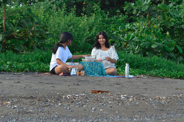 Smiling brightly, two teenage girls sort their belongings and food on a beach mat, enjoying the...