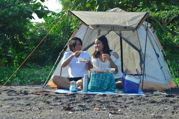 Sitting on a mat under a canopy, two teenage girls share food and drinks while joking, enjoying the...