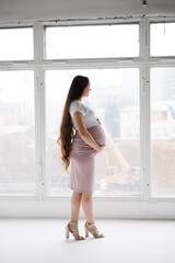 Pregnant woman in dress with very long hair on white background. Dreamy tender girl expecting a child. Portrait of beautiful pregnant woman in white shirt, smile. Window