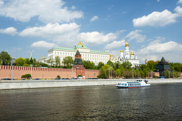 Obraz premium View of the Moscow Kremlin along the Moskva River. The scene includes historic buildings, green trees, and a boat on the water under a blue sky with clouds.