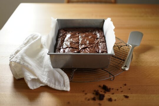 Warm homemade chocolate brownies cooling in a baking pan on a wooden surface with natural light and kitchen utensils nearby.
