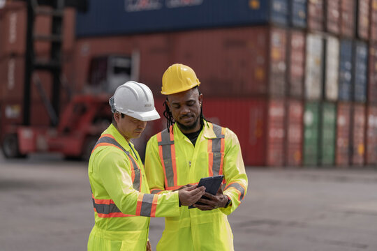 Two logistics workers in high visibility safety gear and hard hats discuss operations at a busy shipping container yard. One is holding a tablet while pointing towards the cargo stacks.