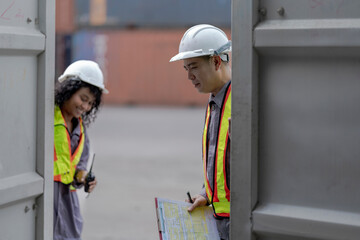Two logistics workers asian man and woman discuss operations at a busy shipping container yard. One is holding a tablet while pointing towards the cargo stacks.