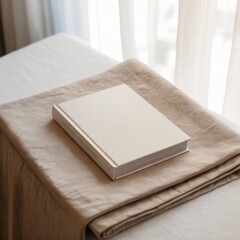 Fototapeta premium A photograph of a book mockup with a clean, minimalist cover design lying on a neatly folded, light beige linen blanket. The book is positioned slightly off-center, showcasing its spine and the subtle