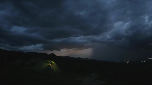 Dramatic zoom in timelapse of couple camping in tent top mountain thunderstorm brews, glowing shelter against dark clouds and lightning in distant horizon.Concept hikers under lightning sky isolated