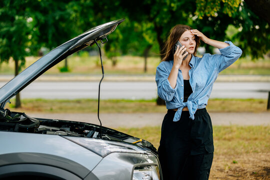 A young woman calls her husband on the phone for help near a broken down new car. Concerned woman explains the problem and waits for help.