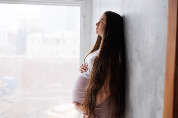 Fototapeta premium Pregnant woman in dress with very long hair on grey background. Dreamy tender girl expecting a child. Portrait of beautiful brunette pregnant woman. Window