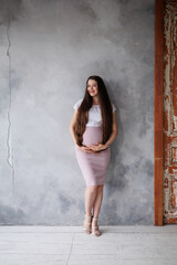 Naklejka premium Pregnant woman in dress with very long hair on grey background. Dreamy tender girl expecting a child. Portrait of beautiful brunette pregnant woman. Window