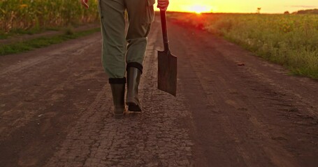 Woman walking with a shovel in hand along a rural dirt road at sunset, wearing green pants and rubber boots.