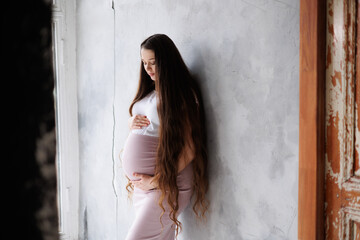 Fototapeta premium Pregnant woman in dress with very long hair on grey background. Dreamy tender girl expecting a child. Portrait of beautiful brunette pregnant woman. Window