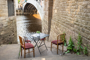 Table and chairs for al fresco dining and drinks seen within the ancient English city of York. The great River Ouse can be seen and the wrought iron bridge to the city.