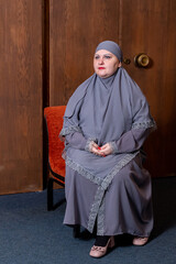 A Muslim woman in a modest gray hijab sits against a wooden door