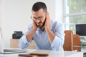 Young businessman suffering from ear pain at table in office
