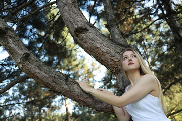 Beautiful young woman enjoys nature while sitting on a tree branch in a sunlit forest during a calm...
