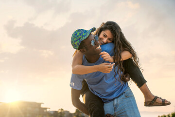 Romantic diverse couple enjoying sunset on the beach, man giving woman a piggyback ride on vacation.