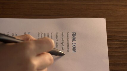 Student’s hand holding a black pen while taking a printed final exam on general knowledge. The paper rests on a wooden desk and the visible section shows the title “FINAL EXAM”