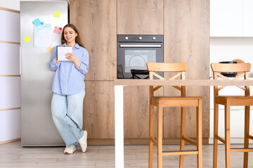 Young woman making to-do list near fridge in kitchen