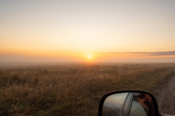 Calm foggy landscape of empty meadow in countryside. Photographer takes photo from car window. Morning with golden light of rising sun