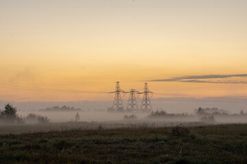 Far away in the field, high-voltage power lines against the orange dawn. Morning fog creeps low over the meadow.
