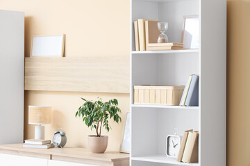 Bookshelf with hourglass and clock near beige wall in room