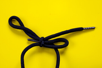 A dark shoelace with metal tips, tied in a decorative knot, lies on a bright yellow background....