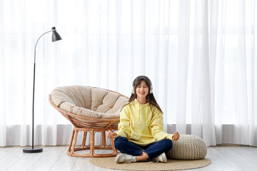 Young woman with headphones meditating on rug near armchair at home