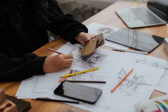 A top down view of a creative workspace where a designer's hands are sketching, surrounded by a laptop, tablet, and blueprints