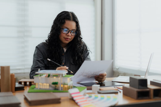 A female architect is reviewing and verifying a technical drawing or blueprint against the physical house model