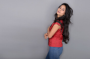 A confident young woman dressed casually in a red top and blue jeans, posing with jovial facial...