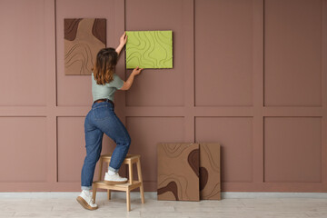 Young woman hanging painting on brown wall, back view