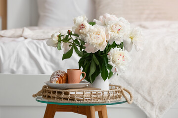 Vase with peony flowers, cup of coffee, croissant and books on table in bedroom