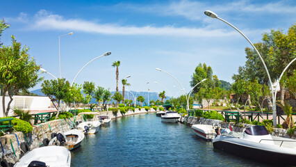Scenic canal with moored boats lined by decorative street lights and lush greenery in Icmeler, Marmaris, Mugla Province, Turkey. Bright sunny day with clear blue sky and distant mountains.