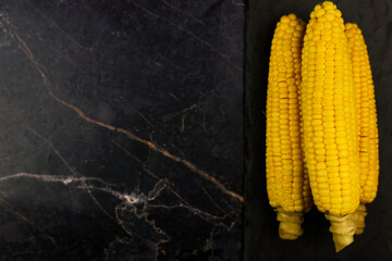 Yellow ripe peeled ears of corn with large grains lie on a black stone plank on the side on a dark background with space for text. The concept of healthy nutrition, natural iron, corn cultivation