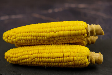 Close-up of yellow, ripe, peeled corn cobs with large grains and stem stems lying horizontally in the center on a dark background. The concept of healthy nutrition, natural iron, corn cultivation