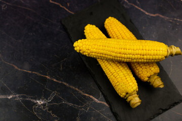 Yellow ripe peeled ears of corn with large grains and stem stem lie on a black stone plank on the side on a dark background with space for text. The concept of healthy nutrition, natural iron, corn