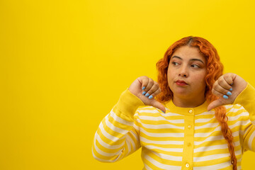 Freckled woman with curly red hair with a disgusted expression with thumbs down on a yellow...